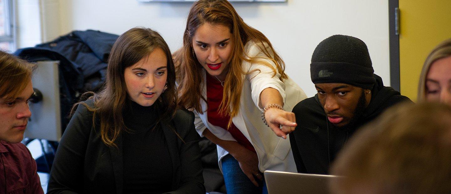 A group of people looking at something on a laptop that one woman is pointing to.